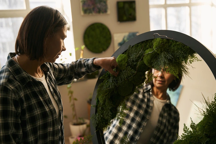Woman Decorating Round Mirror with Greenery A woman carefully attaching green plants to the frame of a large round mirror.