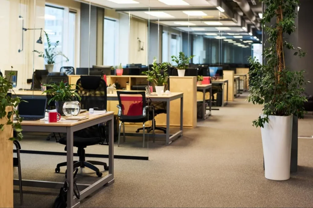 Modern corporate open office with rows of workstations, tall green plants along the aisle, and well-lit clear walkways supporting employee comfort